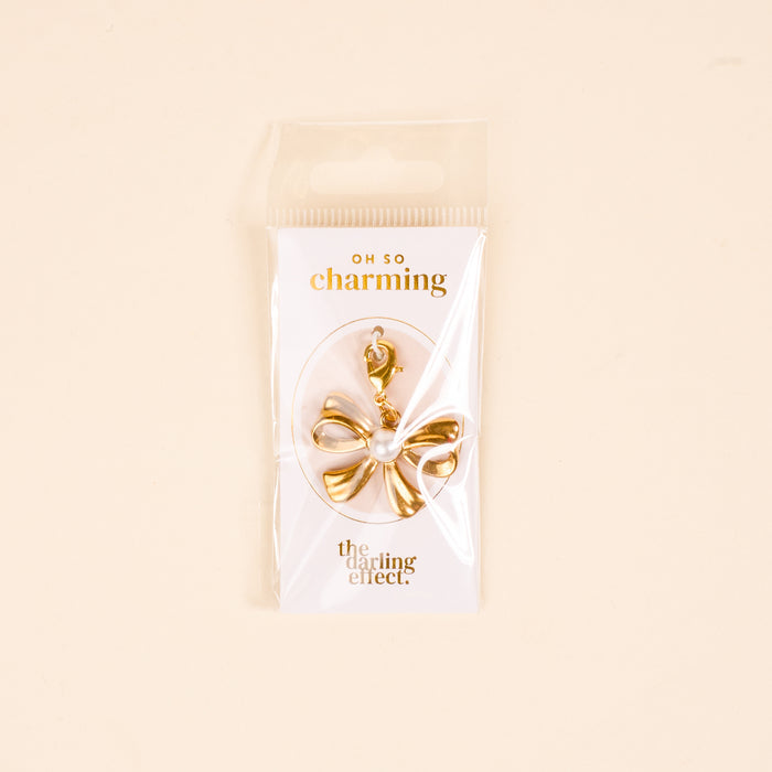The Darling Effect's All Tied Up Jewelry Charm Gold features a gold and white bow shape with a clasp, displayed on an "OH SO charming" card against a light beige background.