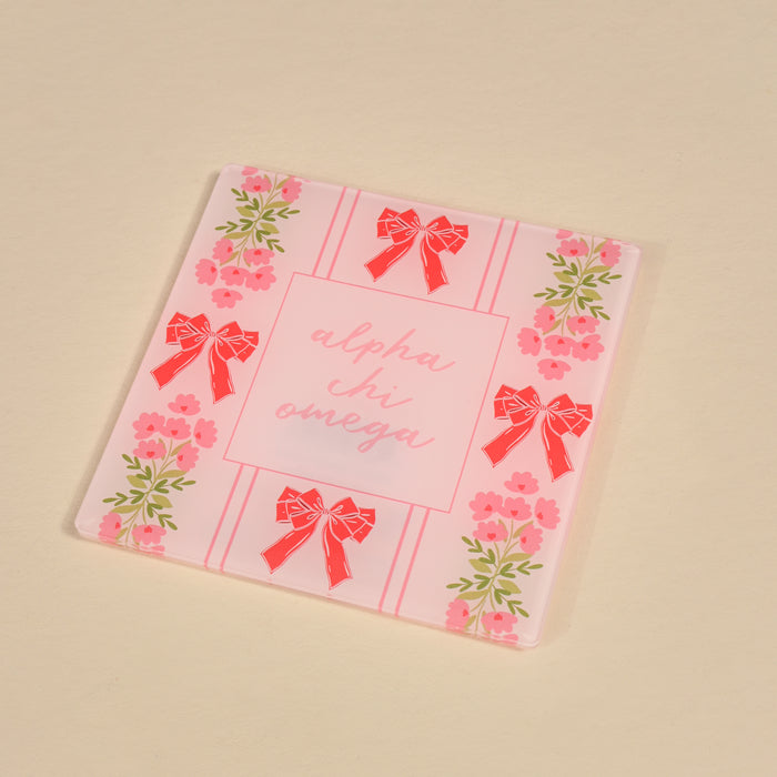 The Darling Effect’s Alpha Chi Omega Acrylic Coaster features pink flowers and red bows on the border with “alpha chi omega” in pink script at the center—a stylish sorority gift or decor for your space.