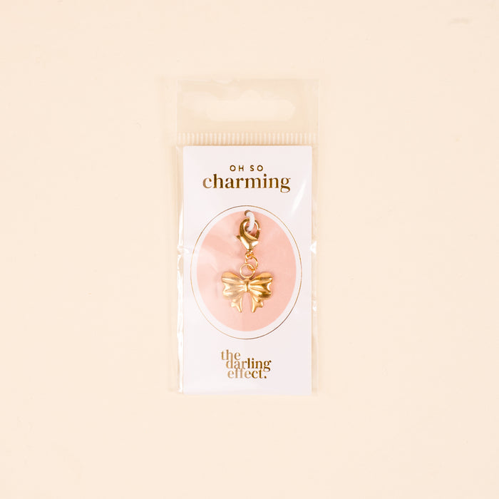 The Bitty Bow Jewelry Charm Gold by the-darling-effect comes attached to a card with โOH SO charmingโ and โthe darling effect.โ, all packaged in a clear plastic bag against a beige backgroundโperfect for necklaces or bracelets.