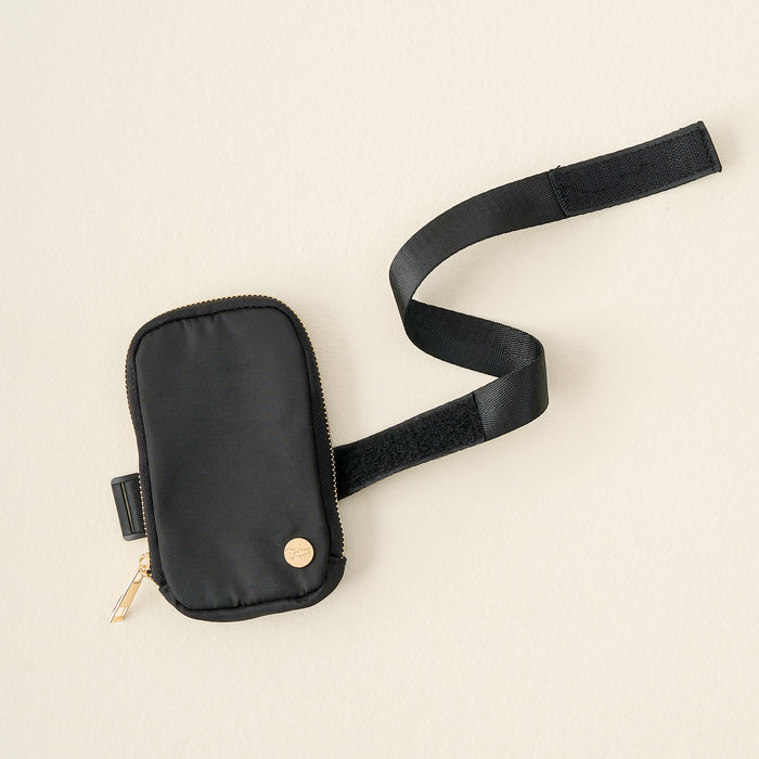 The Darling Effect Black Tumbler Fanny Pack is a small, water-resistant black pouch with a gold zipper and logo, attached to an adjustable Velcro strapβan elegant, stylish tumbler accessory shown on a light beige background.