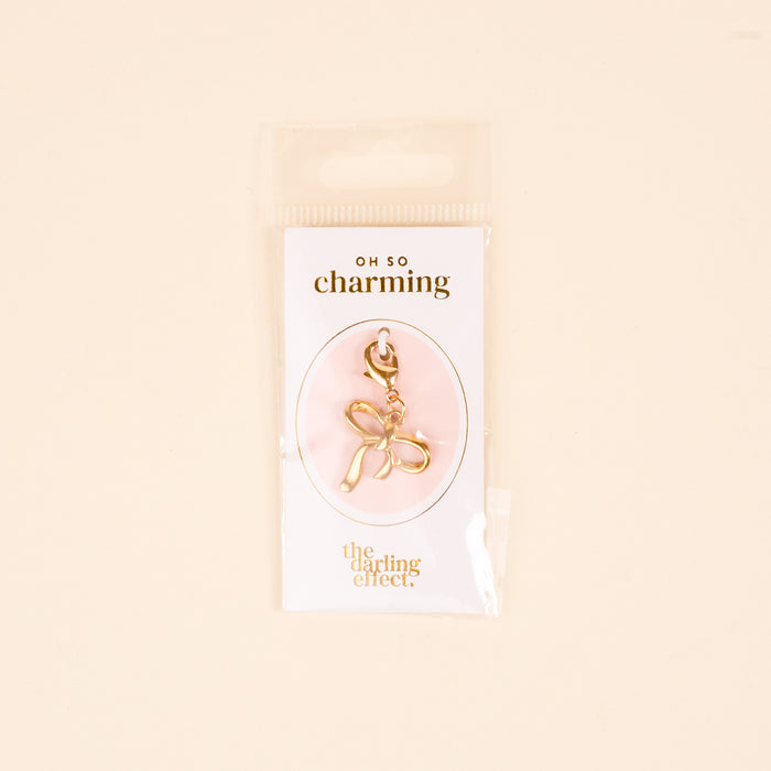 The Bow Jewelry Charm Gold by the-darling-effect is shown in clear packaging on a pink background. The text reads "OH SO charming" and "the darling effect," making it perfect for charm jewelry or cute necklaces.