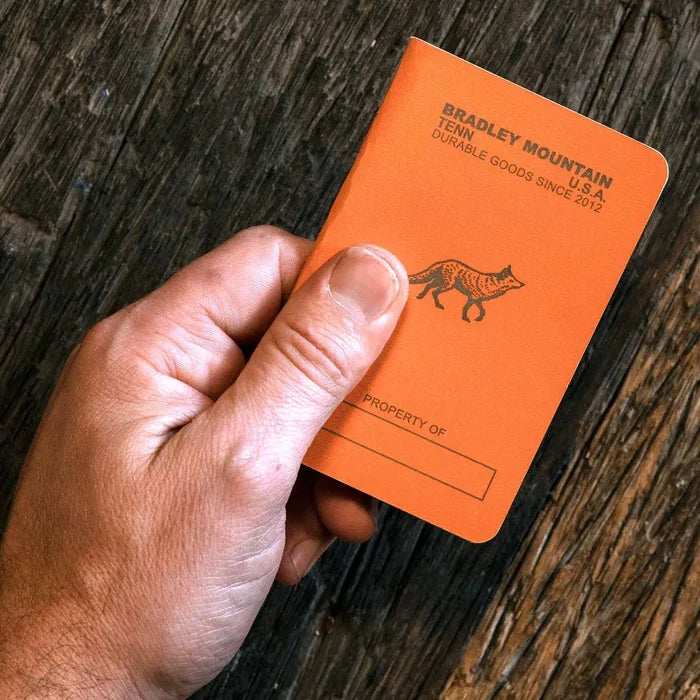 Hand holding Bradley Mountain Pocket Journals 2 Pack in orange on wood table, stylish travel notebook stationery for writers.