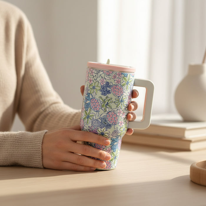 A person in a beige sweater holds The Darling Effect's Coastal Cove 20 oz Lifestyle Flip Straw Tumbler, which features a floral pattern and large handle, while sitting at a light wooden table with books and a white vase behind them.