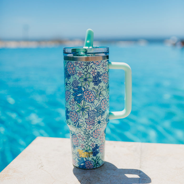The-darling-effect’s Coastal Cove 40 oz Lifestyle Flip Straw Tumbler, featuring a vibrant floral design and handle, rests poolside beside bright blue water under a clear sky.