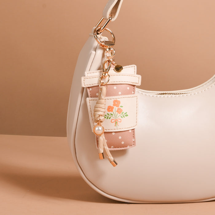A cream-colored handbag is adorned with The Darling Effectβs Coffee Run Vegan Leather Bag Charmβa playful pink floral keychain and a small pouch featuring an orange flower and polka dots, both hanging from a gold clasp on the handle. These charming vegan leather accents elevate the stylish accessory, showcased against a beige background.