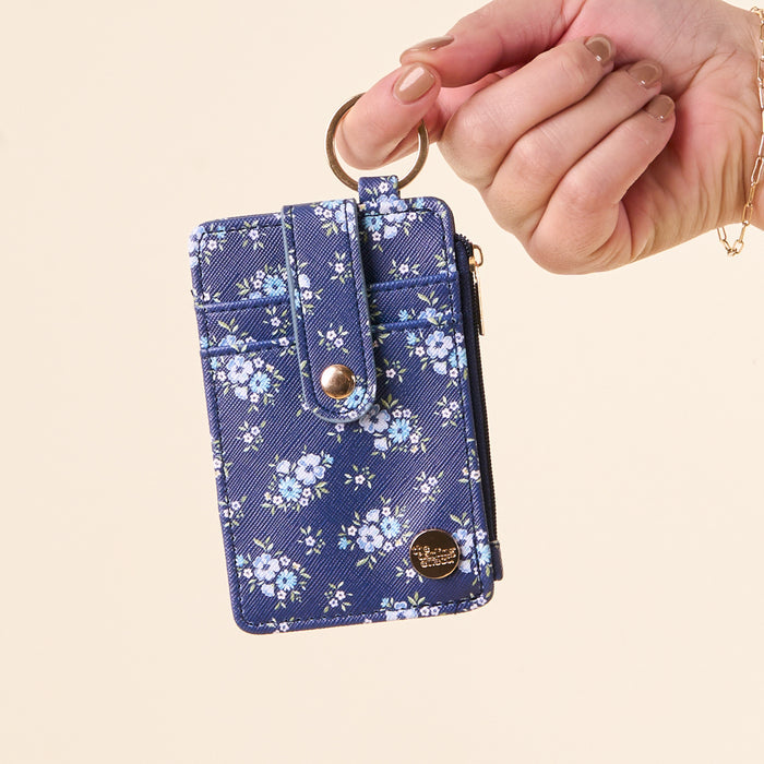 A hand holds the-darling-effect Endless Daydream Navy Keychain Wallet, attached to a small blue zippered coin holder with a white floral pattern, gold button, and side zipper. The background is plain light beige.
