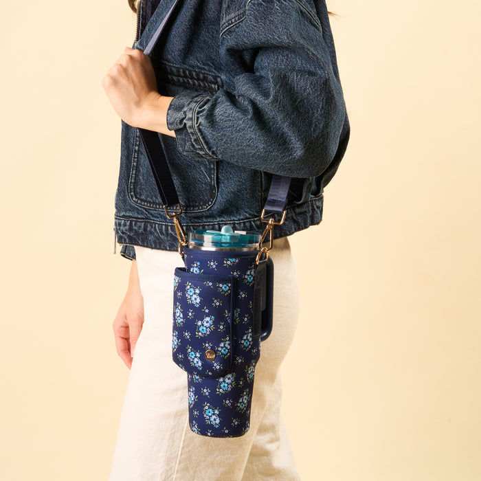 A person in a denim jacket and white pants carries the-darling-effect's Endless Daydream Navy Tumbler Sling, featuring a floral pattern and adjustable strap, for secure hands-free drink carrying. The background is plain light beige.