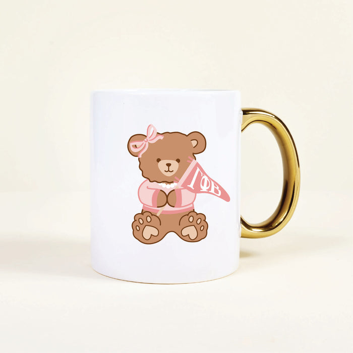 Enjoy your morning coffee with The Darling Effect's Gamma Phi Beta 11 oz Cozy Cute Ceramic Mug, featuring a brown teddy bear in a pink sweater holding a Gamma Phi Beta pennantโperfect for sorority members or as an adorable gift.