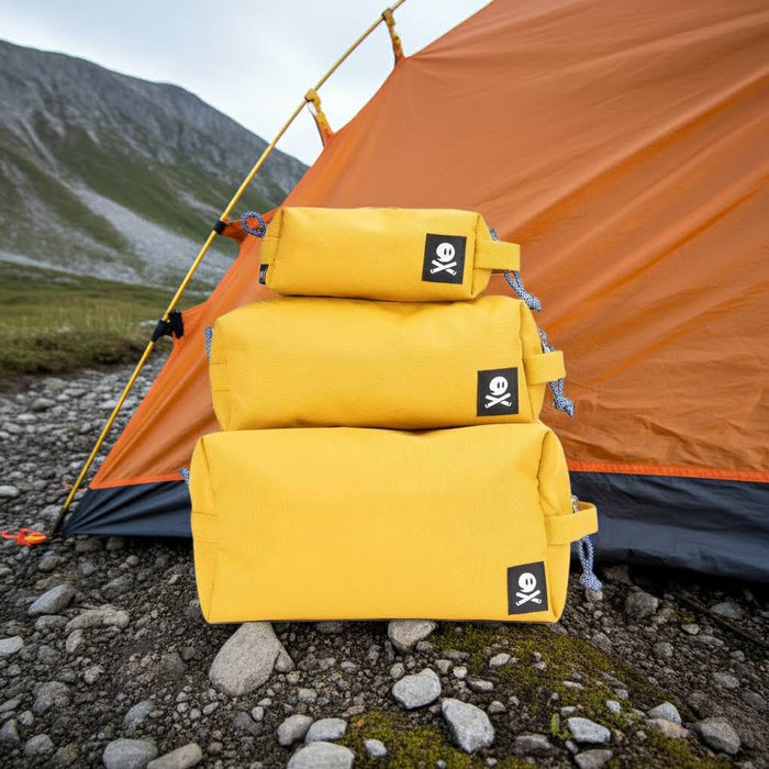 Three Ghost Outdoors yellow Dopp kits stacked by an orange tent on rocky groundโcamping travel toiletry bags outdoors.