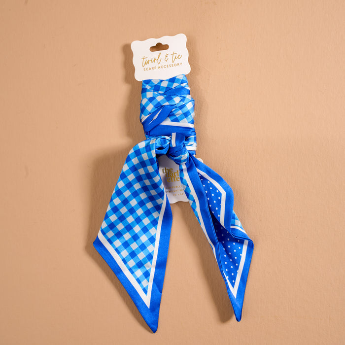 The Gingham Blue Twirl & Tie Scarf Accessory by The Darling Effect features a chic combination of blue and white checks with playful polka dots in lustrous satin, neatly folded and tied against a beige background. Its attached product tag highlights the endless styling options this versatile accessory offers for any occasion.