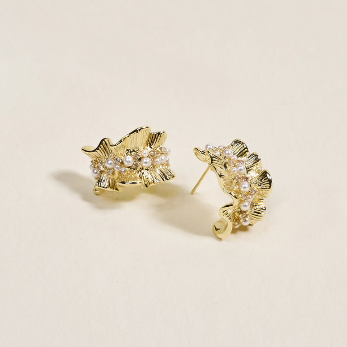 The Darling Effect's Golden Ruffle Standout Earrings feature a high-shine finish and pearl accents along the center, displayed on a light beige background.