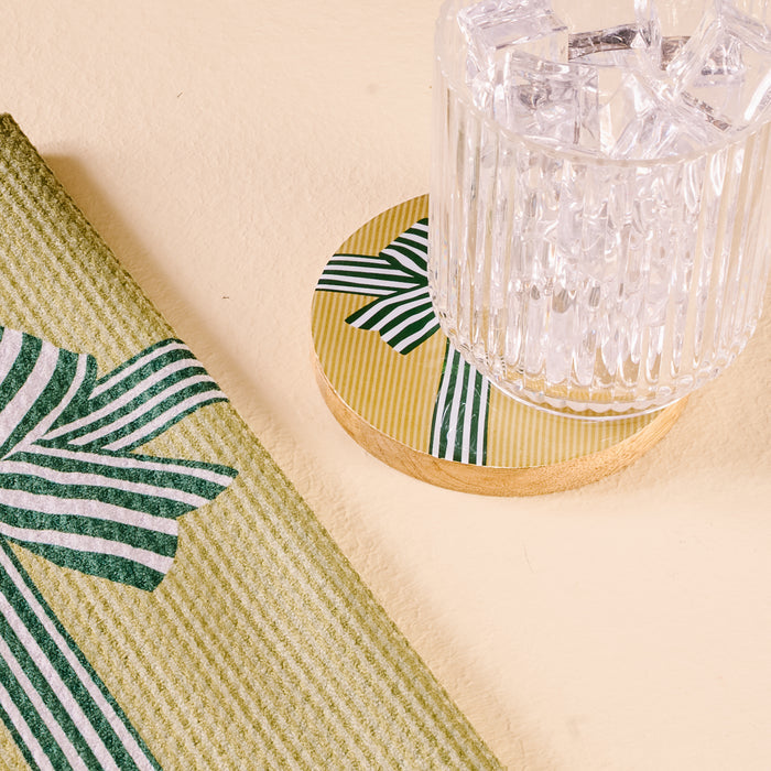 A clear glass with ice sits on The Darling Effect Holiday Gift Bow Green Wood Enamel Coaster, showcasing yellow stripes and green ribbon accents. Next to it is a folded green napkin with a coordinating ribbon design.