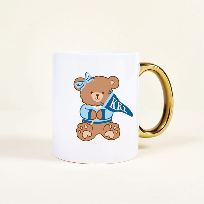 The Darling Effectโs Kappa Kappa Gamma 11 oz Cozy Cute Ceramic Mug features a white base, gold handle, and a brown teddy bear in blue clothes with Greek letters ฮฮฮโperfect for any Kappa Kappa Gamma member.