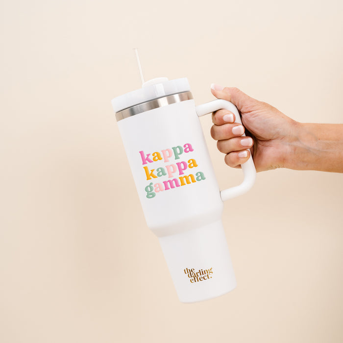 A hand holds The Darling Effect Kappa Kappa Gamma 40 oz Take Me Everywhere white tumbler with a straw, featuring colorful sorority text and The Darling Effect’s gold logo, set against a pale beige background.