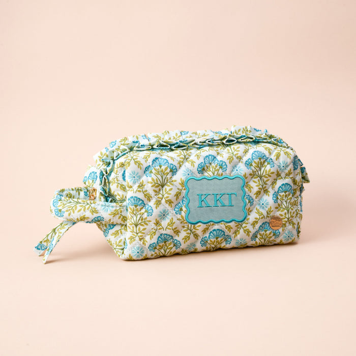 The Darling Effect’s Kappa Kappa Gamma Essentials Quilted Cosmetic Pouch features a floral design, zipper, side handle, and embroidered KKG on a light blue label—ideal for storing sorority must-haves on a pale peach background.