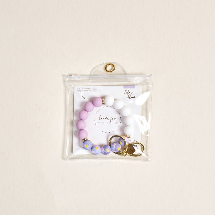 The Lilac Bloom Hands-Free Keychain Wristlet by the-darling-effect features pink, white, and blue silicone beads with a gold keyring and clasp, all in a clear zippered pouch with a branded card inside.