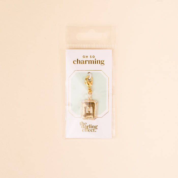The M Jewelry Charm Initial Letter Gold by the-darling-effect features a gold-plated charm attached to a lobster clasp, displayed on white Oh So Charming packagingβan ideal gift for personalized jewelry lovers.
