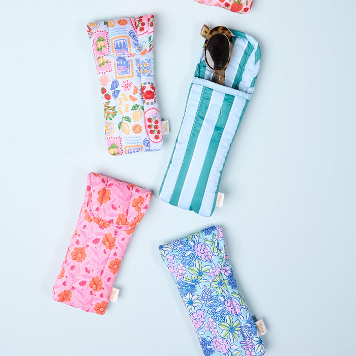 Five vibrant, machine-washable eyeglass cases with floral and patterned prints are displayed on a light blue surface. The Darling Effect's Oh So Fancy Pink Puffy Sunglasses Case holds a pair of sunglasses and features a plush pink design.