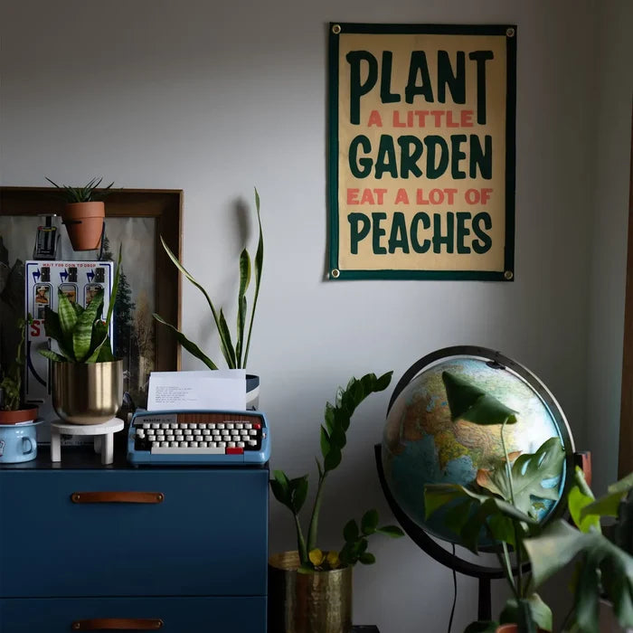 The Oxford Pennant x John Prine banner displayed over dresser with potted plants and vintage globe, stylish home decor.