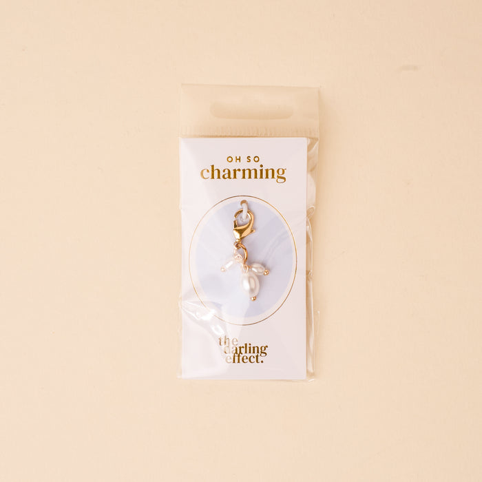 The Pearl Trio Jewelry Charm Gold by the-darling-effect comes in a clear pouch on a card with "OH SO charming" and "the darling effect." The soft beige background makes it perfect for any charm bar collection.