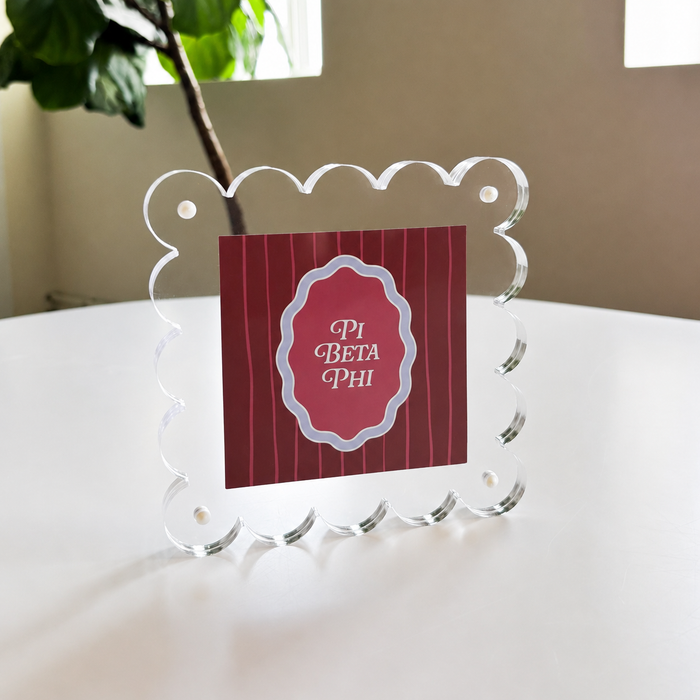 The Darling Effect's Pi Beta Phi Square Scalloped Acrylic Frame displays a red PI BETA PHI card on a white table, with leafy plants and windows behindโperfect for showcasing memories in modern style.