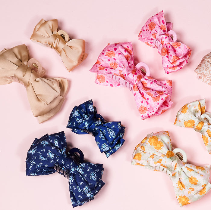 The Darling Effect’s Poppin' Petals Hot Pink Mini Satin Bow Claw Clips, featuring floral prints and vibrant colors like beige, blue, pink, and white, are displayed on a light pink background.