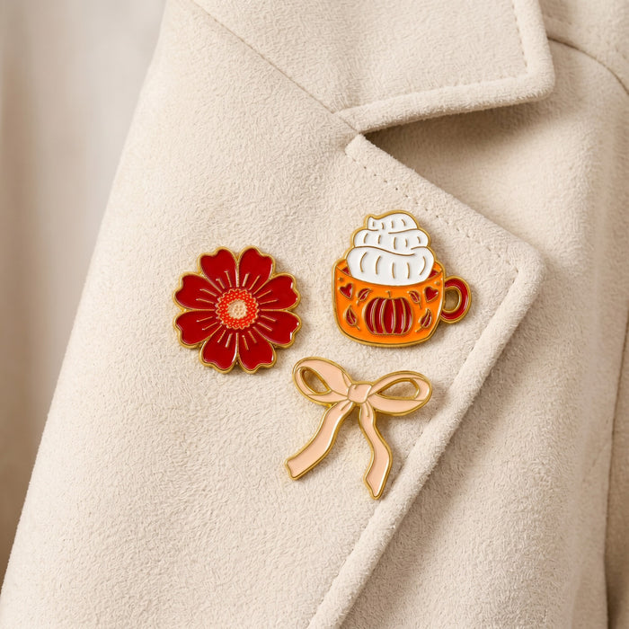 Pumpkin Spice The Pin Pack by The Darling Effect features three collectible pinsβa red flower, an orange pumpkin mug topped with whipped cream, and a light gold ribbon bowβthat add a touch of personality and charm to any outfit.