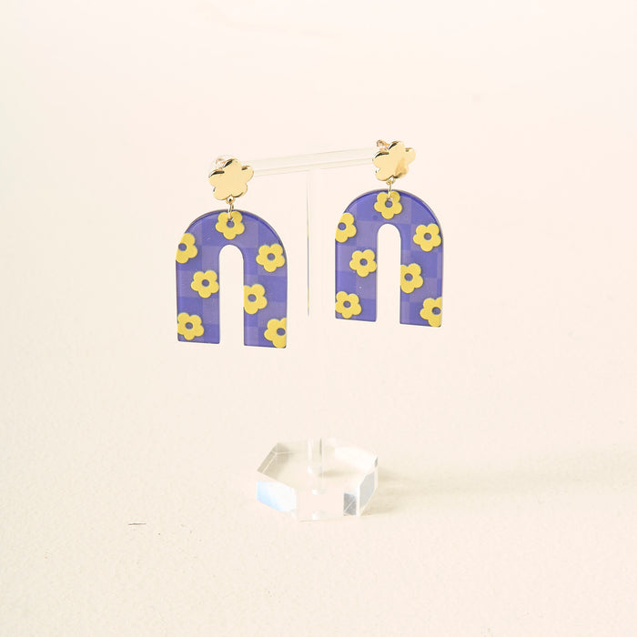 Show your school spirit with The Darling Effect's Purple Yellow Game Day Acrylic Earrings—arch-shaped with yellow flower patterns on a purple background, hanging from gold cloud studs, and displayed on a clear stand.