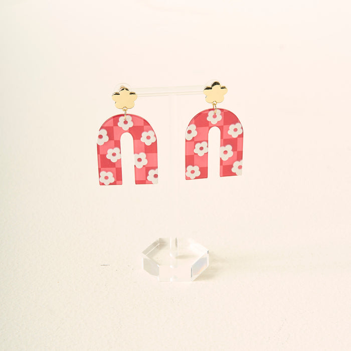 A pair of red, arch-shaped acrylic earrings with white flower patterns hang from gold-colored flower-shaped studs, displayed on a clear stand—Red Game Day Acrylic Earrings by The Darling Effect, perfect for showing your school spirit on game day.