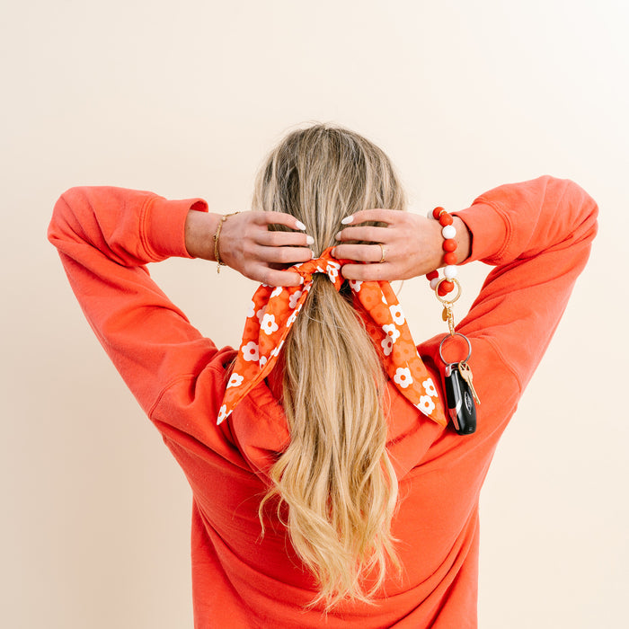 A woman with long blonde hair, seen from behind, wears an orange sweatshirt and ties her hair with a red Game Day Hair Scarf with Scrunchie by The Darling Effectβan ideal team spirit accessory. She also has a bracelet and a keychain with keys on her left wrist.