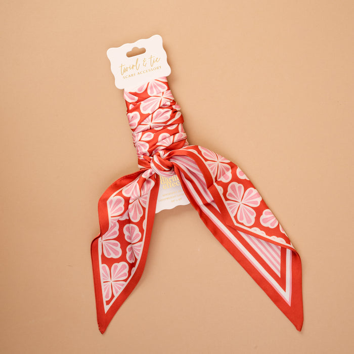 The Sienna Spice Twirl & Tie Scarf Accessory by The Darling Effect is a satin scarf in an eye-catching red and pink floral pattern, elegantly displayed against a beige backdrop. Featuring the brandβs signature label, a central knot, and pointed ends, this versatile accessory offers endless styling possibilities.