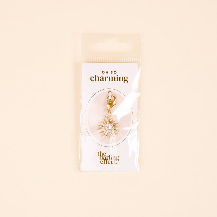 The Star Burst Jewelry Charm Gold by the-darling-effect features a gold star with clear gems on a lobster clasp, perfect for your charm bar. Packaged on a white card with OH SO charming and the-darling-effect logosโtimeless elegance for your collection.