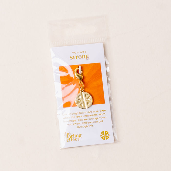 The Strong Jewelry Trait Charm Gold by the-darling-effect is a gold keychain with a circular strength charm, packaged on a card that says โYou are strongโ with an encouraging message and displays the the-darling-effect logo at the bottom.