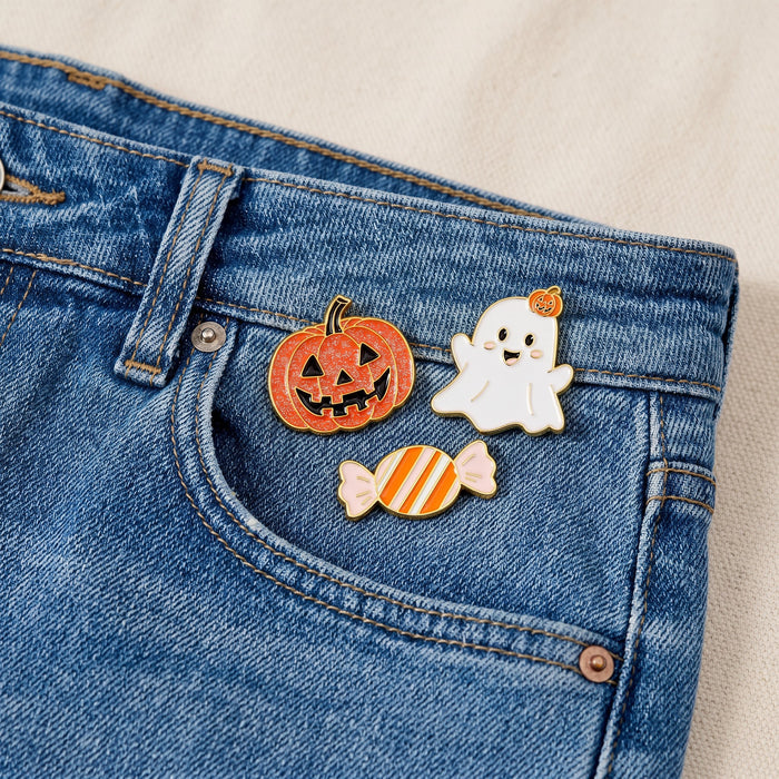 Three collectible pins from The Darling Effectβs Trick or Treat The Pin Packβa jack-o-lantern, a smiling ghost, and a striped candyβare attached to the small pocket of blue denim jeans. These expressive accessories stand out against the light-colored fabric background.