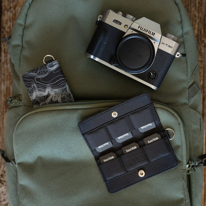 Green backpack, camera, patterned keychain, and Wildtree Topographic Map SD/Switch Card Holder on wooden surface.