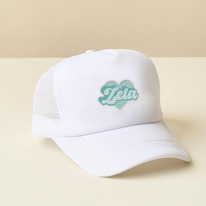 The Darling Effect Zeta Tau Alpha White Trucker Hat features white mesh, a curved brim, green heart patch, and cursive "Zeta" embroidery. Perfect sorority gift, shown on a light surface.