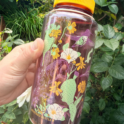 Hand holds 32oz Mustard Beetle Nalgene Anza Borrego bottle with floral, hummingbird design and bright yellow lid.