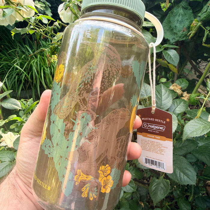 A hand grips a 32oz Mustard Beetle Nalgene Water Bottle by Joshua Tree, outdoors among lush green plants and desert scenery.