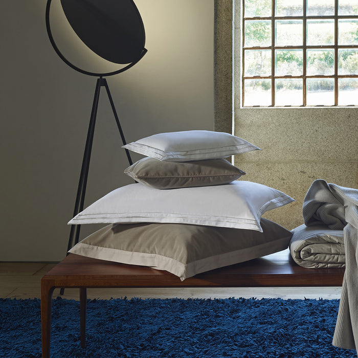 Stack of Alba white sand pillowcase sham on a wooden bench with a window and lamp in the background