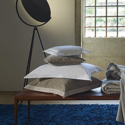Stack of Alba white sand pillowcase sham on a wooden bench with a window and lamp in the background