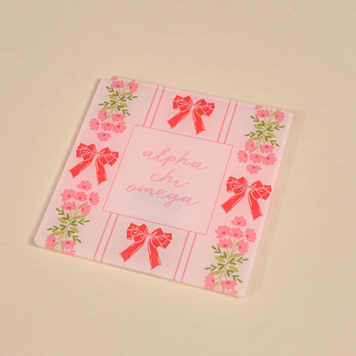 The Darling Effect’s Alpha Chi Omega Acrylic Coaster features pink flowers and red bows on the border with “alpha chi omega” in pink script at the center—a stylish sorority gift or decor for your space.