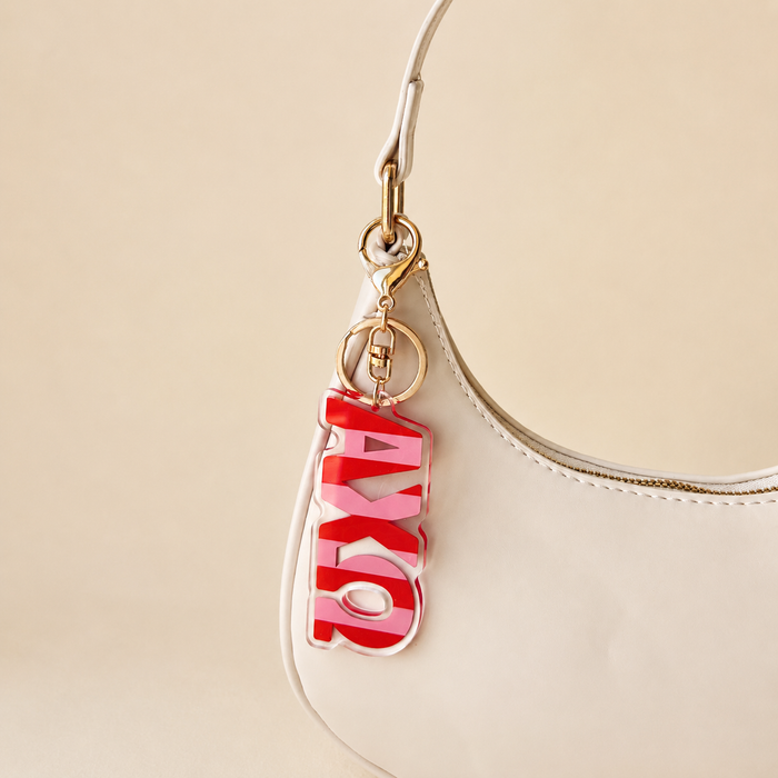 A white handbag with gold hardware features The Darling Effect’s Alpha Chi Omega Stripes Acrylic Keychain on the handle—an ideal, stylish gift for sorority members or a memorable bid day accessory.
