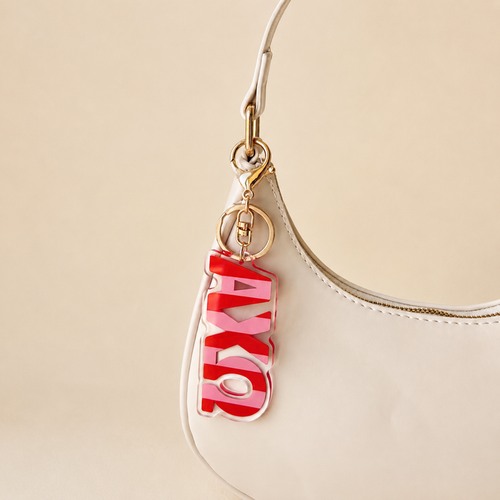 A white handbag with gold hardware features The Darling Effect’s Alpha Chi Omega Stripes Acrylic Keychain on the handle—an ideal, stylish gift for sorority members or a memorable bid day accessory.