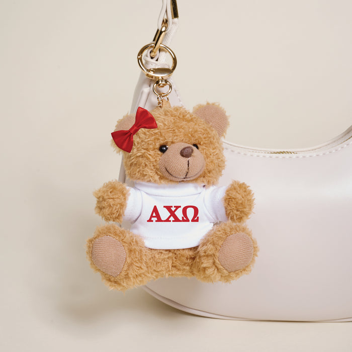 The Darling Effect Alpha Chi Omega Teddy Bear Bag Charm features a small bear with a red bow and white shirt displaying red AXΩ letters, hanging from a keychain—perfect for accessorizing any white handbag.
