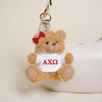 The Darling Effect Alpha Chi Omega Teddy Bear Bag Charm features a small bear with a red bow and white shirt displaying red AXΩ letters, hanging from a keychain—perfect for accessorizing any white handbag.