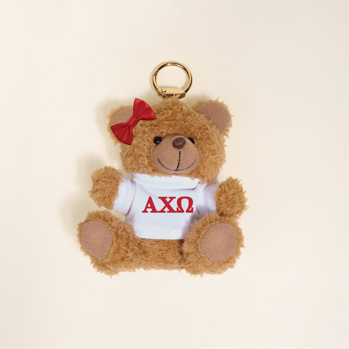 The Darling Effect’s Alpha Chi Omega Teddy Bear Bag Charm is a small brown bear keychain with a red bow and white shirt displaying red ΑΧΩ Greek letters, shown on a light beige background.