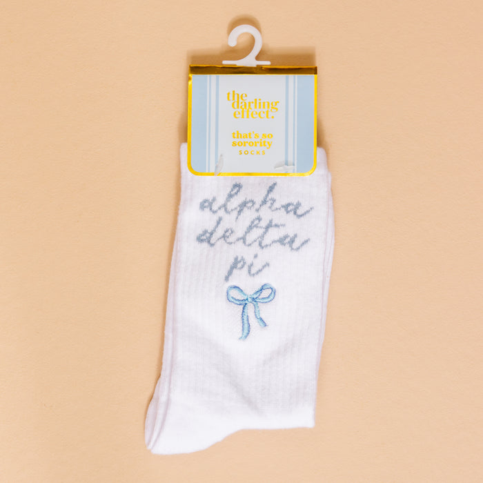 Show off your sorority spirit with The Darling Effect's Alpha Delta Pi Embroidered Crew Socks—white socks featuring blue script, a cute blue bow, and a yellow "the darling effect thats so sorority socks" label, displayed on a beige background.