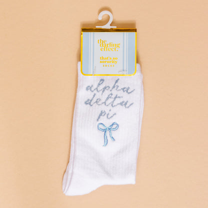 Show off your sorority spirit with The Darling Effect's Alpha Delta Pi Embroidered Crew Socks—white socks featuring blue script, a cute blue bow, and a yellow "the darling effect thats so sorority socks" label, displayed on a beige background.
