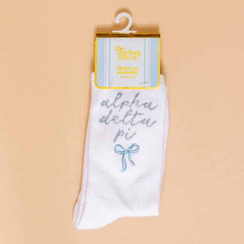 Show off your sorority spirit with The Darling Effect's Alpha Delta Pi Embroidered Crew Socks—white socks featuring blue script, a cute blue bow, and a yellow "the darling effect thats so sorority socks" label, displayed on a beige background.