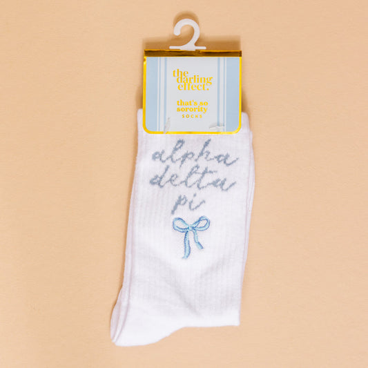 Show off your sorority spirit with The Darling Effect's Alpha Delta Pi Embroidered Crew Socks—white socks featuring blue script, a cute blue bow, and a yellow "the darling effect thats so sorority socks" label, displayed on a beige background.
